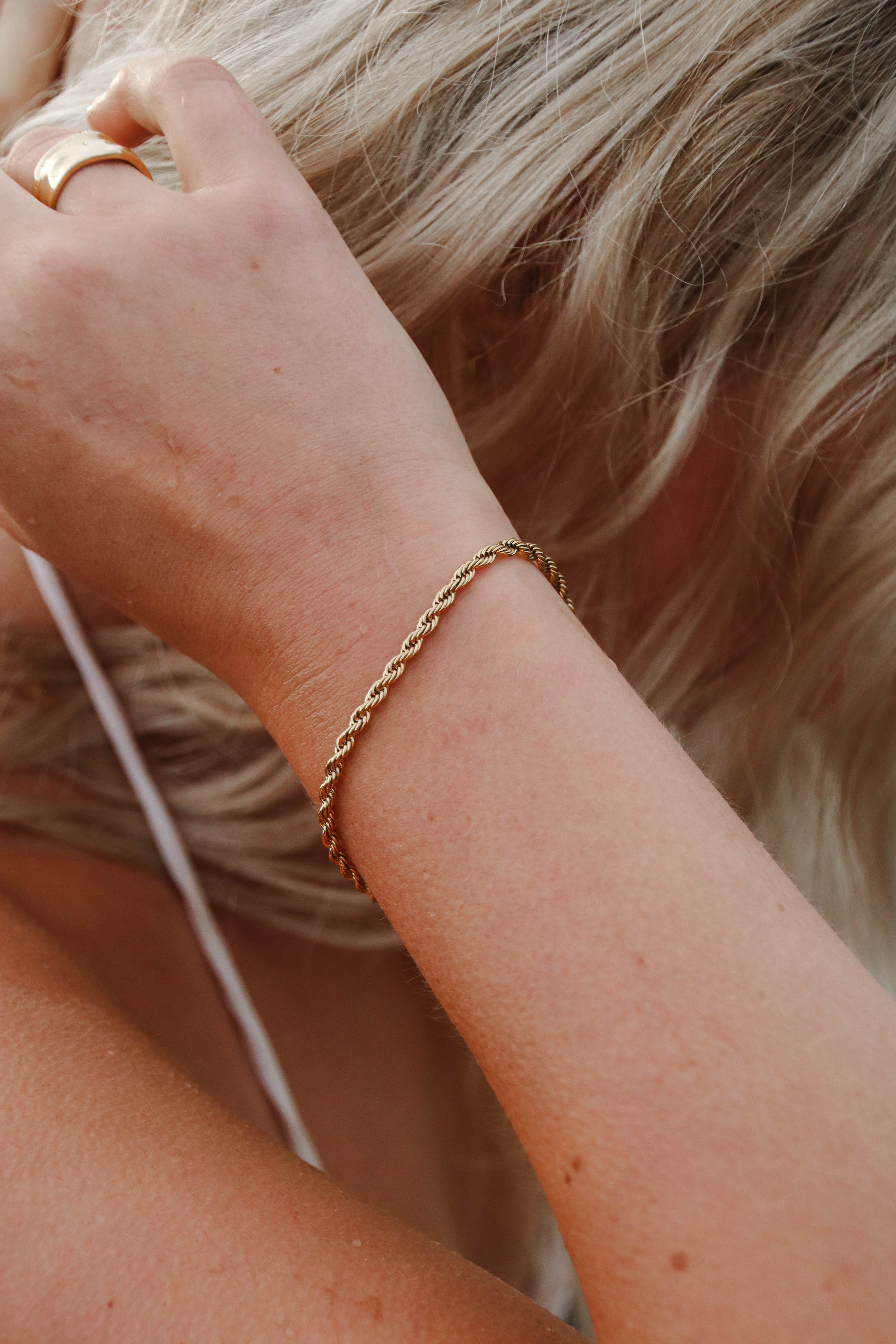 Närbild på en handled med Aloe Allie Bracelet – ett elegant, tvinnat armband i guld som fångar ljuset vackert. Armbandet bärs tillsammans med en chunky ring och blondt hår i bakgrunden, för en avslappnad men lyxig känsla.