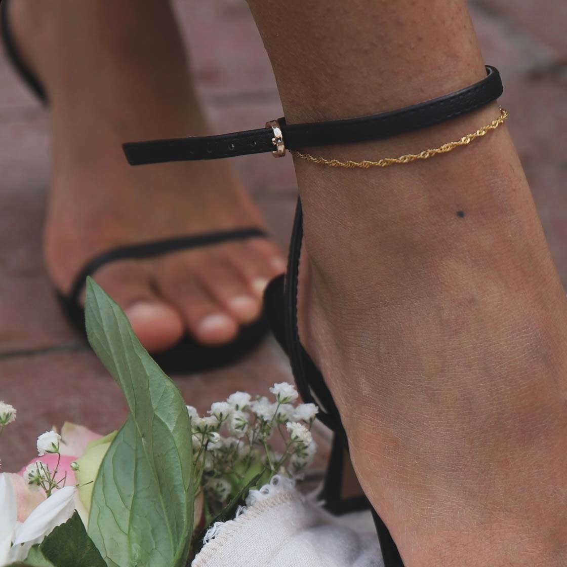 Siri Singapore Anklet från Alora Avenue – tunn, guldfärgad fotlänk med tvinnad design. Bärs tillsammans med svarta klackar och omgiven av blomdetaljer – en elegant och vattentålig accessoar för sommarens alla tillfällen.