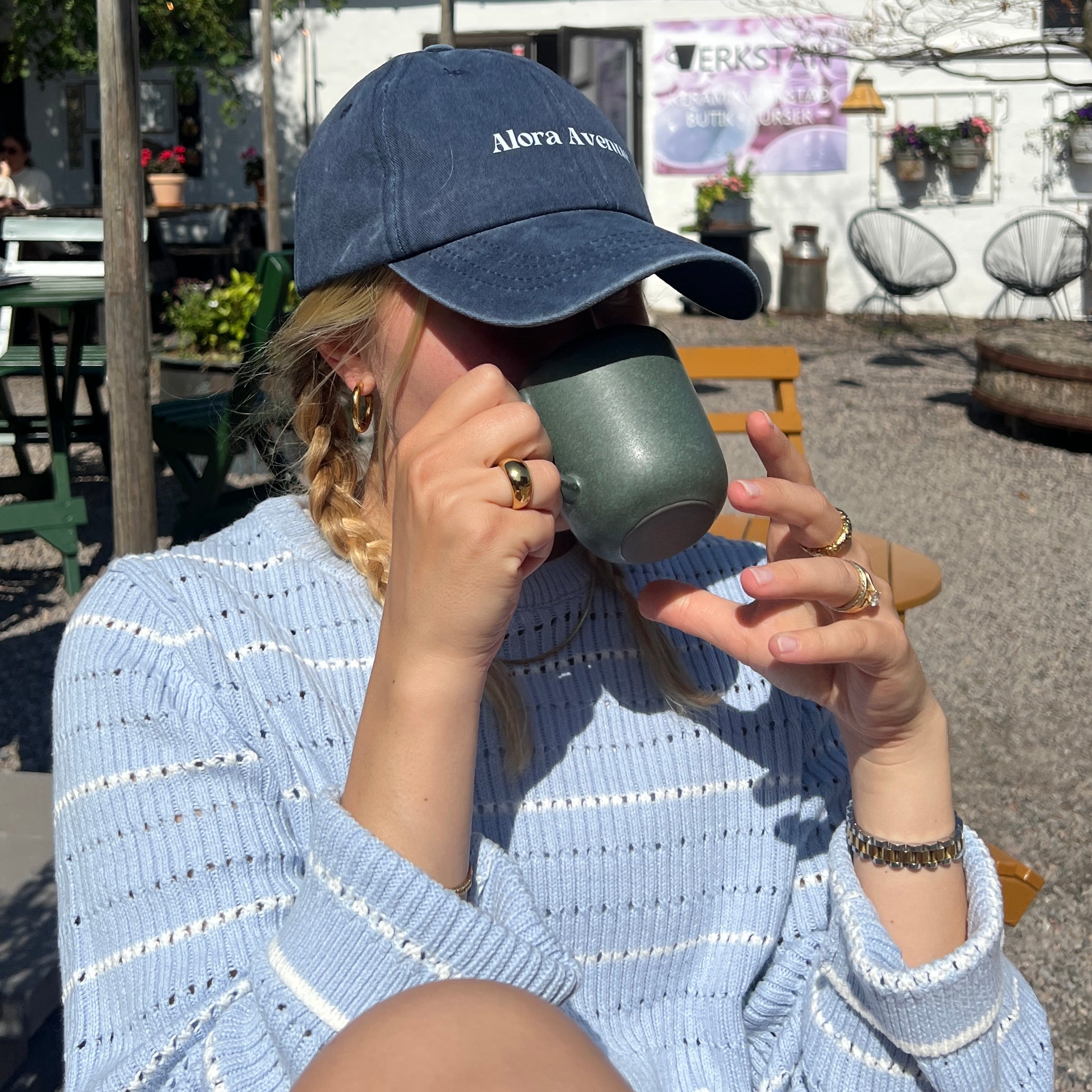 Kvinna i blå stickad tröja och marinblå Alora Avenue-keps, avnjuter kaffe i solen – avslappnad vardagsstil med guldsmycken och somrig känsla.