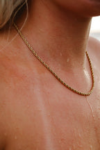 Aloe Annie Necklace – ett tvinnat guldfärgat halsband – vilar mot en solbränd, vattendroppstäckt hud efter ett dopp i havet. Sval, somrig känsla med närbild på halsen.