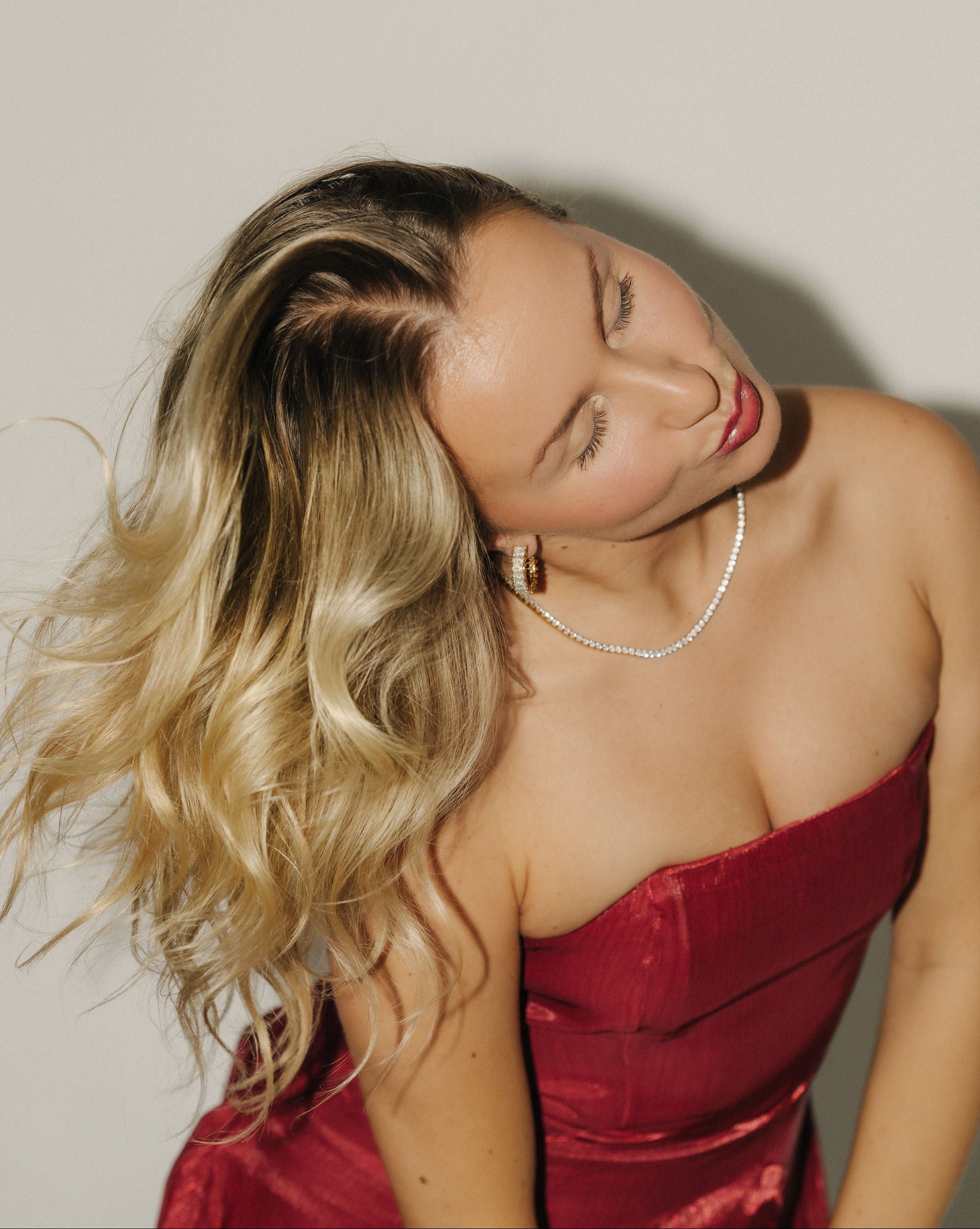 Kvinna i röd klänning bär Alora Avenue Amara Tennis Necklace och guldfärgade hoops. Smyckena är vattentåliga, allergivänliga och tillverkade i rostfritt stål med 18K PVD-plätering. Perfekta som julklapp till henne eller som eleganta accessoarer till julfest och nyår.