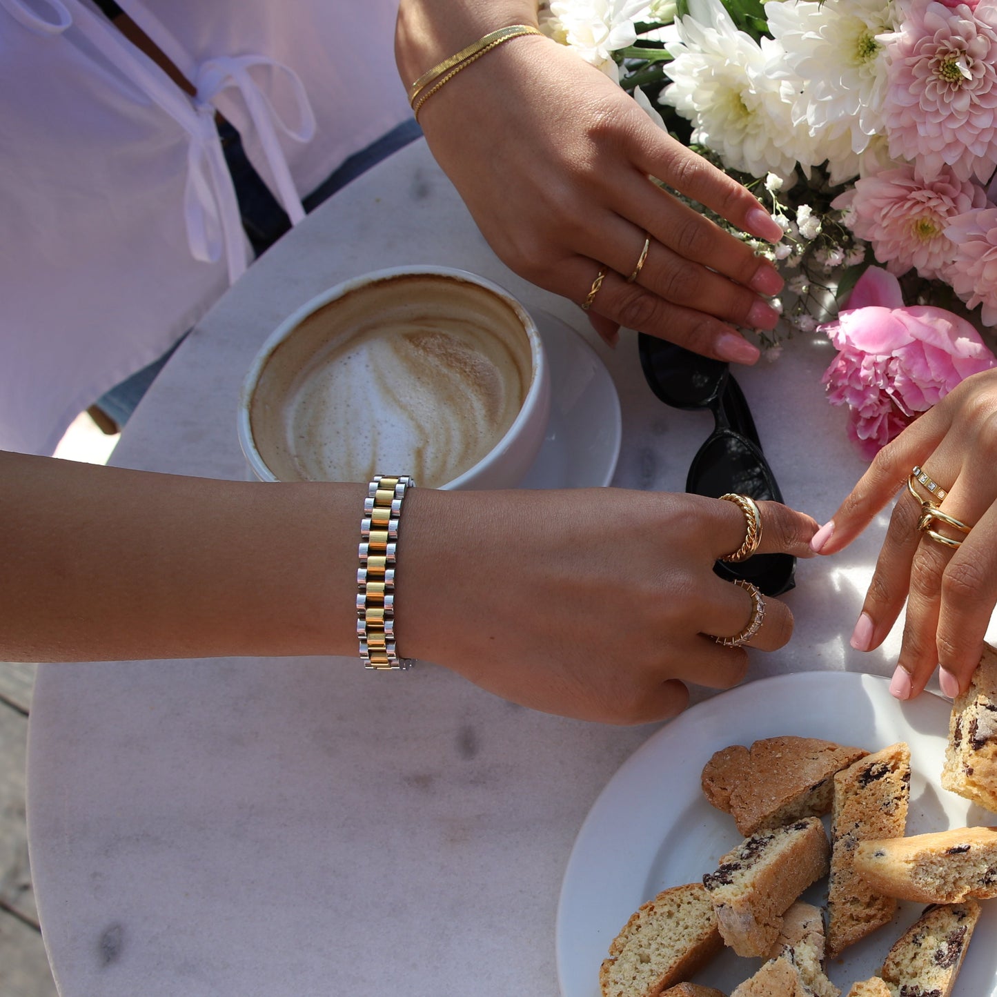 Analog Ava-armband i guld och silver syns på en kvinna som njuter av en solig fikastund med kaffe. Armbandet har en bred, klockinspirerad design utan urtavla, kombinerar modern elegans med exklusiv stil och matchar andra guldaccessoarer. Tillverkat av allergivänligt, rostfritt stål med hållbar PVD-plätering – ett tidlöst, vattentåligt och sofistikerat smycke för alla tillfällen.