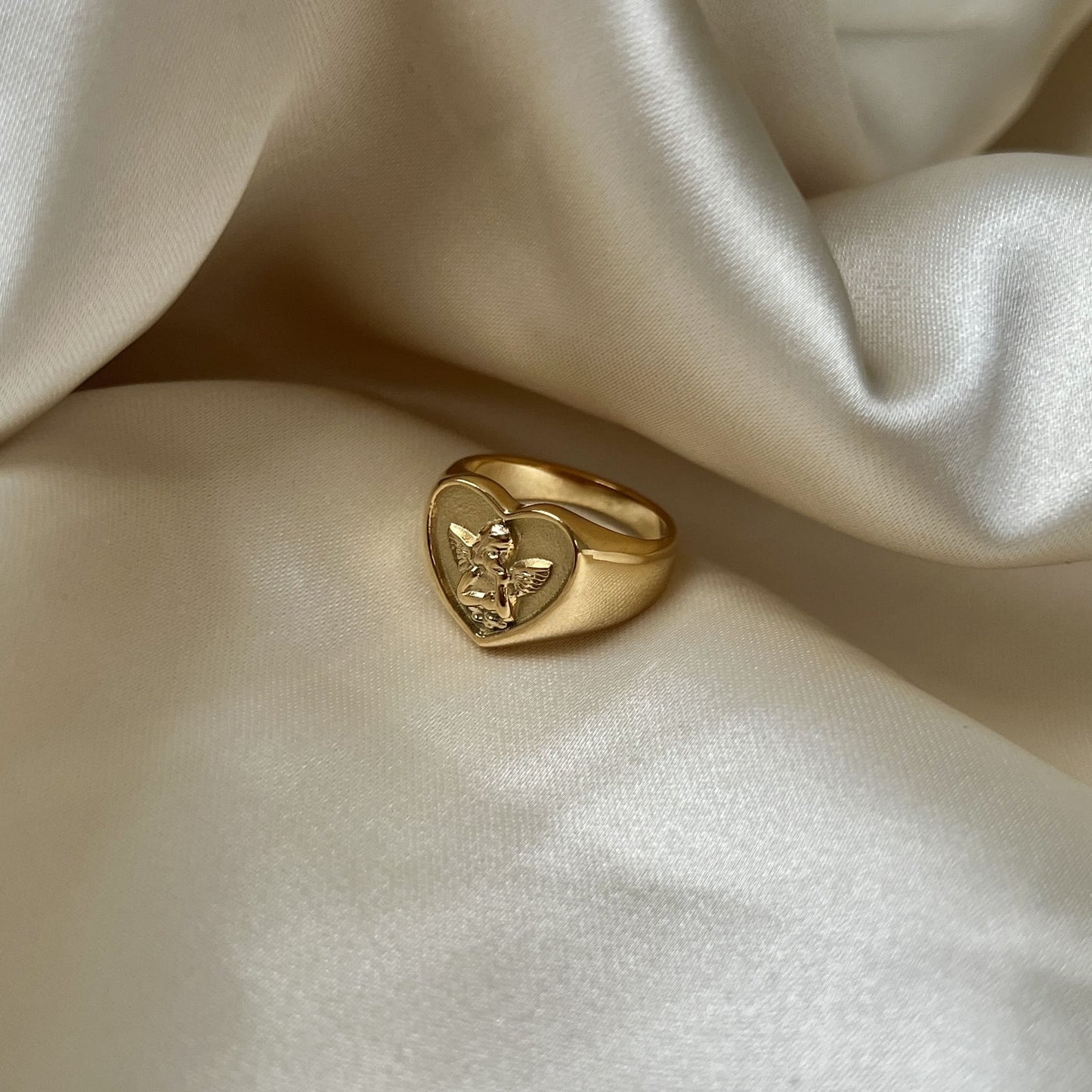 Angelic Love Ring i guld bärs på ett finger av en kvinna som fikar i solskenet, omgiven av kakor och andra guldsmycken. En tidlös accessoar som utstrålar kärlek och stil.