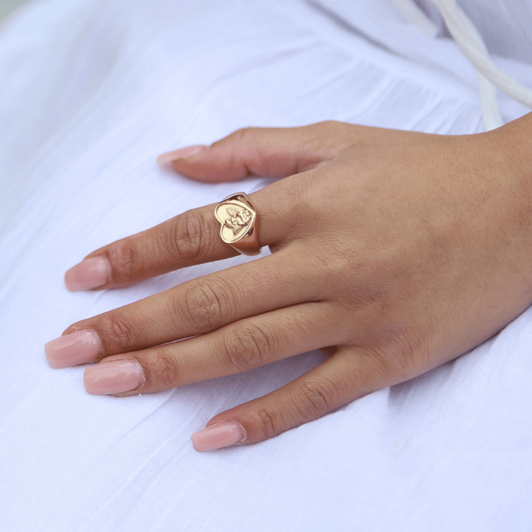 Angelic Love Ring, en vacker guldring med ett hjärta och ängeldetalj, bärs på ett finger som vilar avslappnat mot ett knä – en elegant accessoar för alla tillfällen.