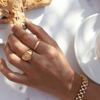Angelic Love Ring i guld bärs på ett finger av en kvinna som fikar i solskenet, omgiven av kakor och andra guldsmycken. En tidlös accessoar som utstrålar kärlek och stil.