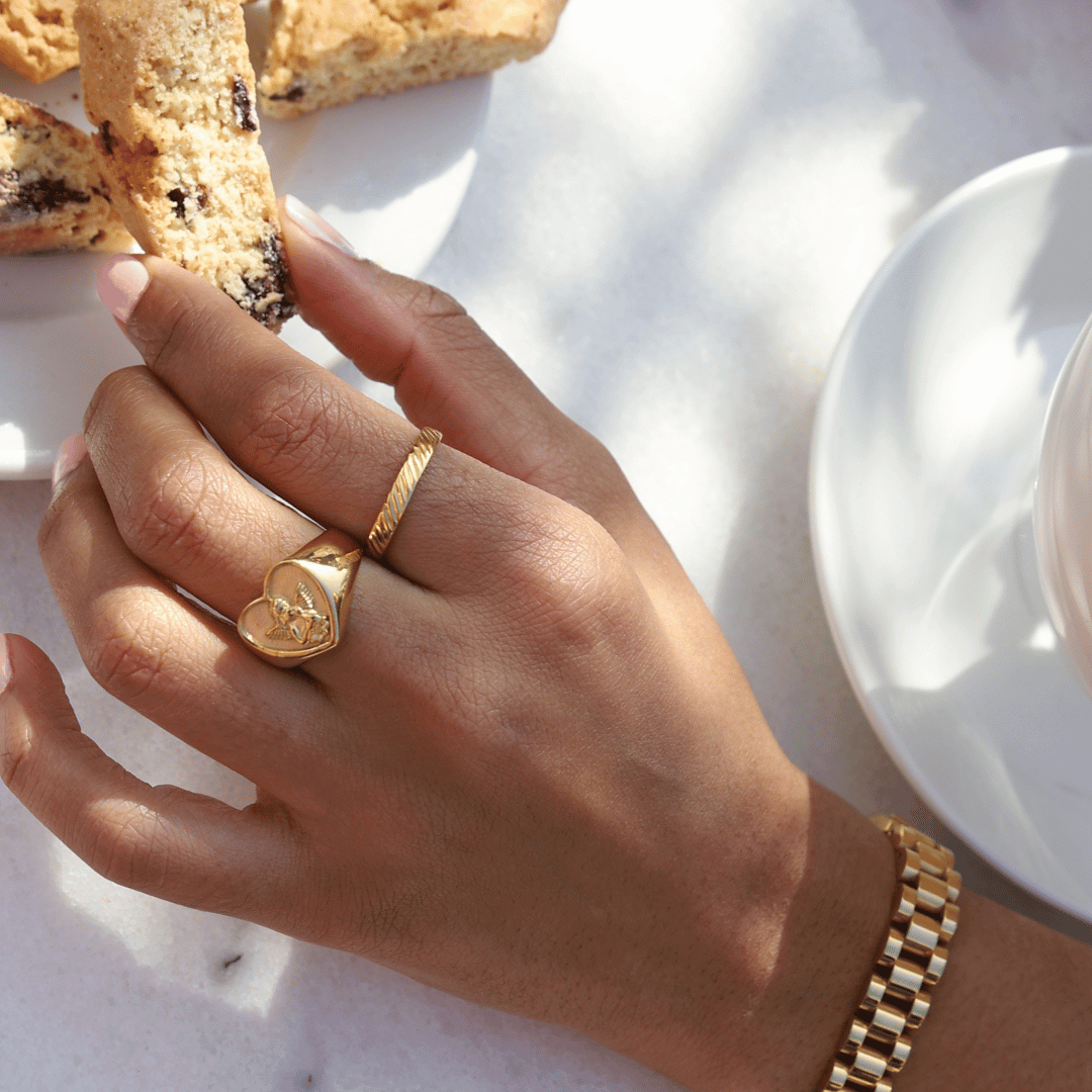 Angelic Love Ring i guld bärs på ett finger av en kvinna som fikar i solskenet, omgiven av kakor och andra guldsmycken. En tidlös accessoar som utstrålar kärlek och stil.