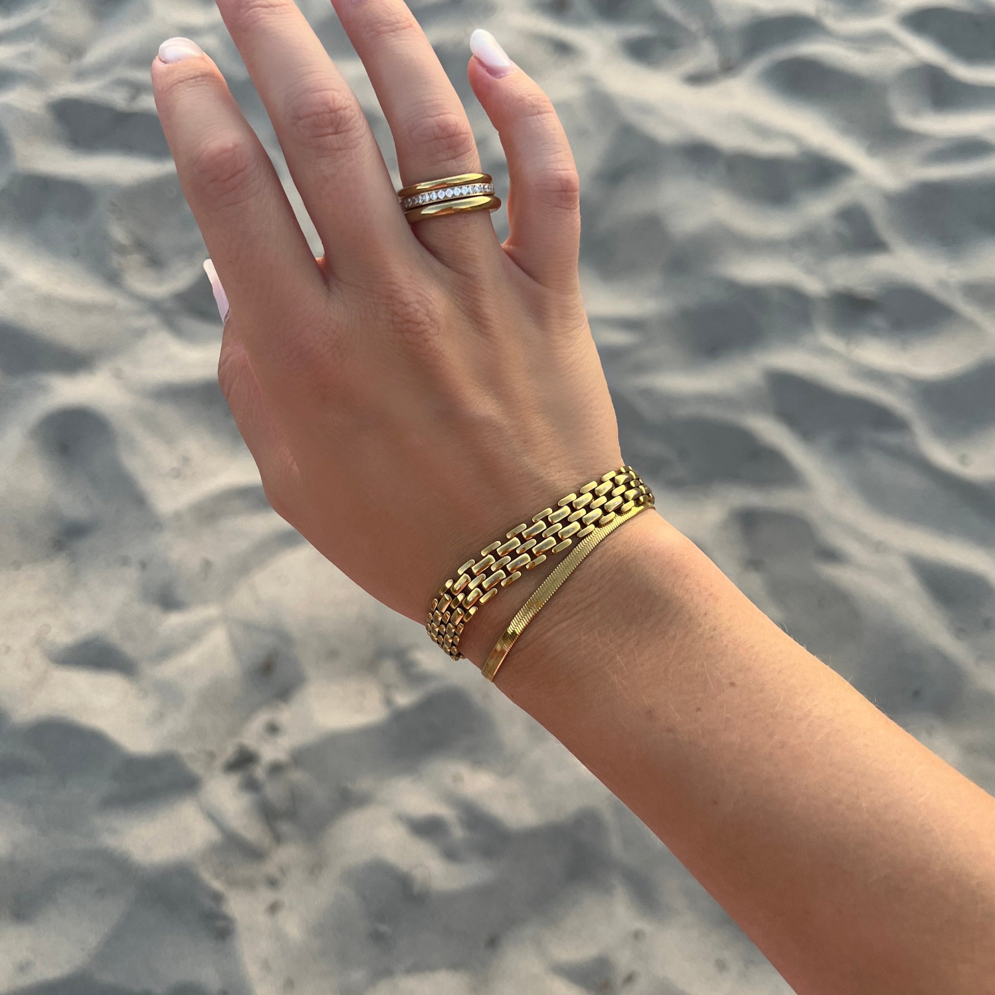 Chloe Cobra Bracelet på kvinnas arm, elegant snake-kedja i guld, som glänser i solen vid en vacker strand och havet.