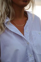 Clara Cobra Necklace – ett glansigt, platt guldfärgat halsband – bärs till en vit skjorta med öppen krage. Smycket reflekterar ljuset elegant mot huden.