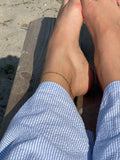 Siri Singapore Anklet från Alora Avenue – elegant guldfärgad vristkedja i singaporekedja. Fotograferad på fot vid stranden med blårandig sommarskjorta. Vattentåligt smycke i rostfritt stål med 18K PVD-guldplätering.