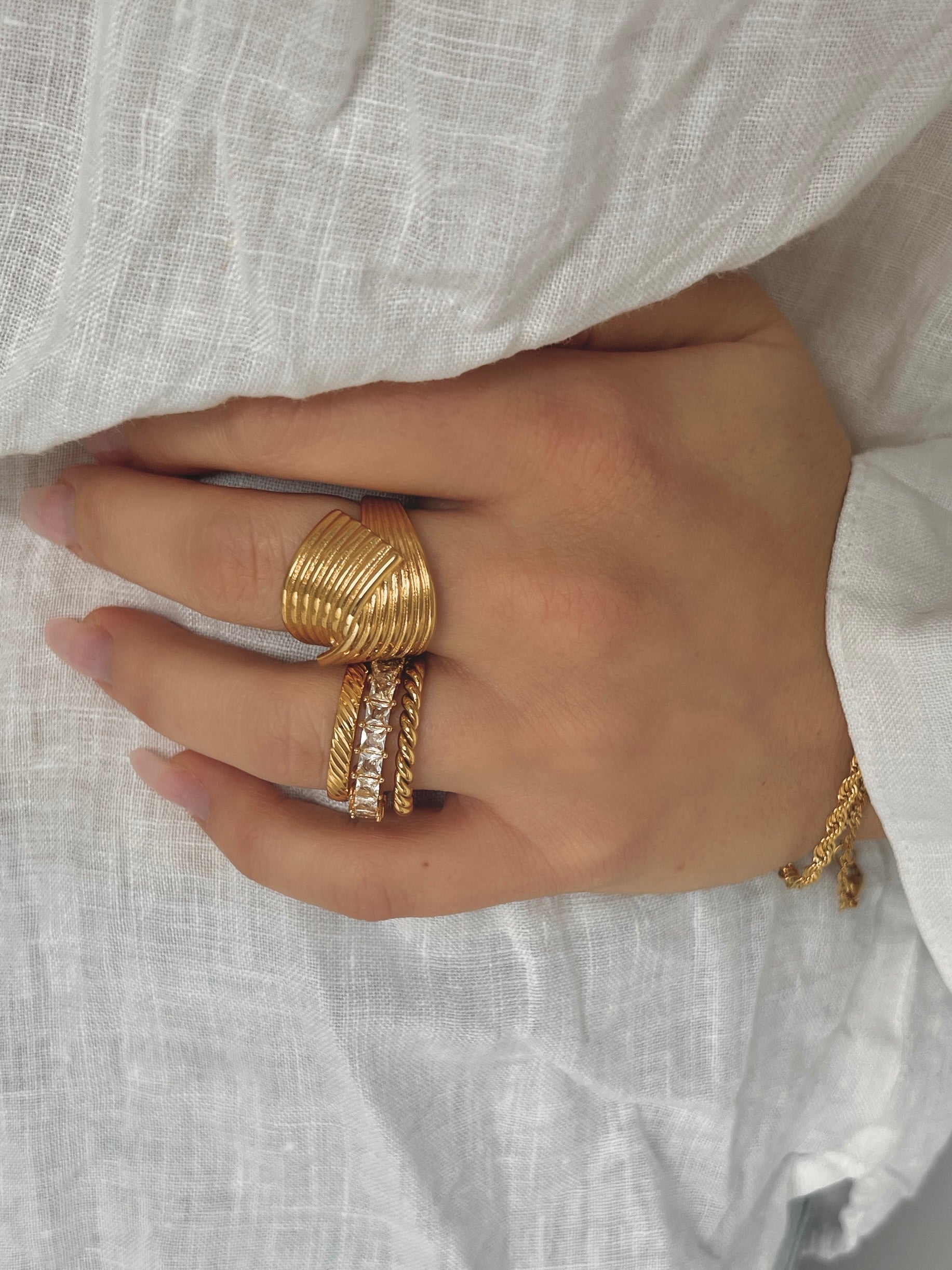 Hedda Huggie Ring från Alora Avenue – guldig bred ring med räfflad design. Fotad på handen mot vit linneskjorta tillsammans med andra stapelringar.
