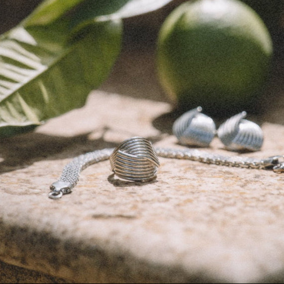 Närbild på Hedwig Huggie Ring från Alora Avenue i silver, fotograferad i varmt solljus på kalkstensyta bredvid gröna citrusfrukter. En vattentålig och allergivänlig ring i rostfritt stål med skulptural form – perfekt för dig som vill bära stil med karaktär varje dag.