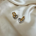 Lydia Love Loops från Alora Avenue – örhängen i tvåtonad design med guldfärgade hoops och hjärtformade hängen i blank silverfinish. Vattentåliga och allergivänliga smycken i rostfritt stål, fotograferade på satin – perfekta som present eller glammig vardagsdetalj med känsla.