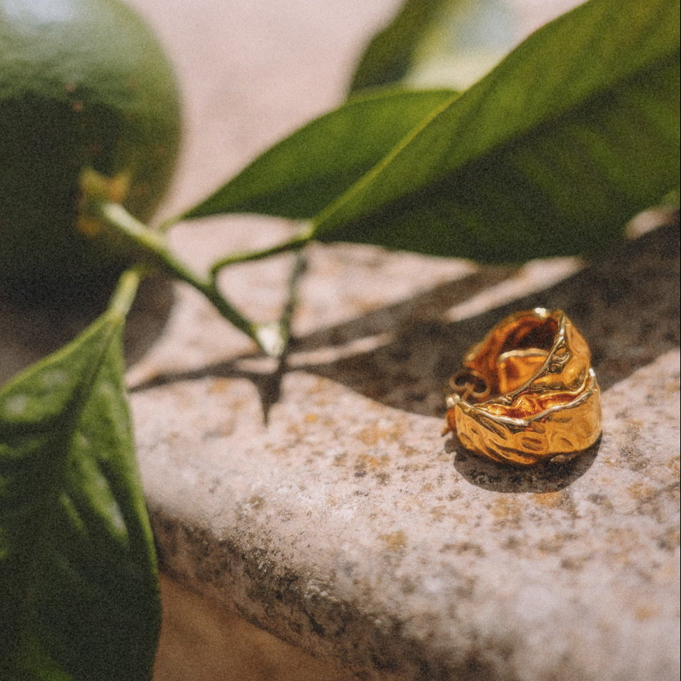 Marigold Chunky Hoops från Alora Avenue i varm solglans, liggande på kalksten intill citrusfrukt och blad. Vattentåliga och allergivänliga guldörhängen i rostfritt stål med organisk form – för dig som vill bära ett statement med själ, varje dag.