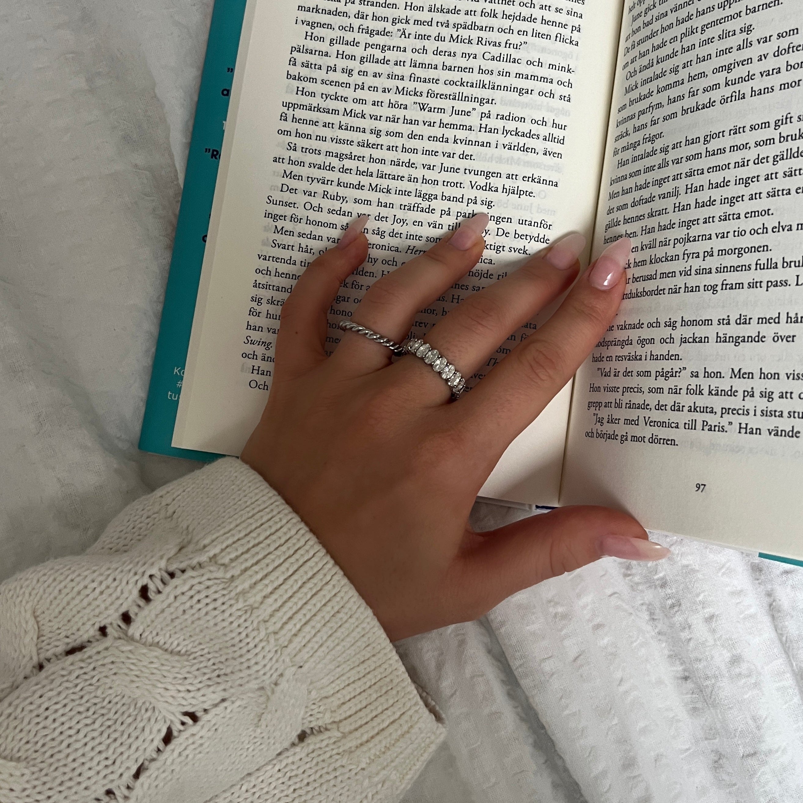 Hand som håller i en bok medan solen skymtar över ett vitt täcke – prydd med Alora Avenues snurrade silverring Milly Moon Ring och en glittrande stenring. En stilla stund med stil.