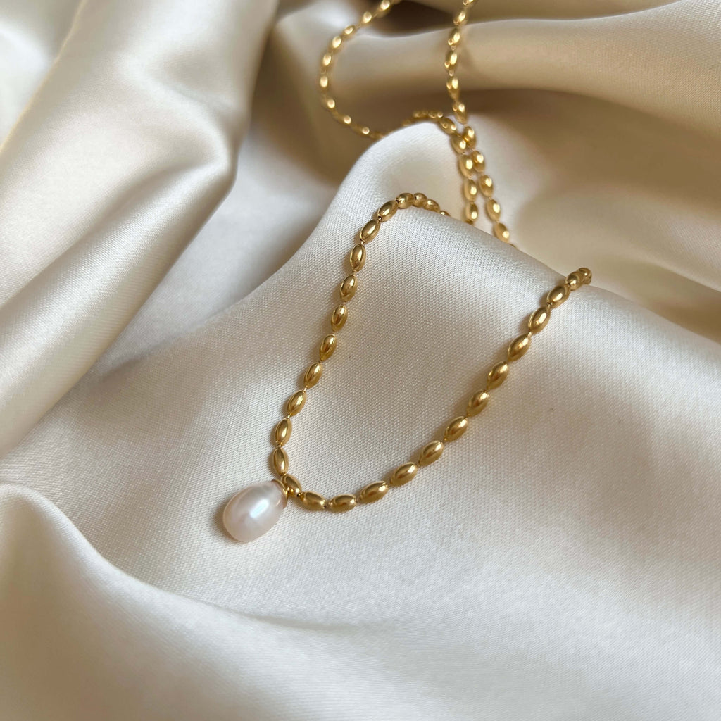 Polly Pearl Necklace från Alora Avenue i närbild – ett vattentåligt halsband i guldfärgat rostfritt stål med droppformad sötvattenspärla, liggande på cremevitt siden. Ett allergivänligt smycke som kombinerar klassisk elegans med modern hållbarhet – perfekt som vardagssmycke eller gåva.