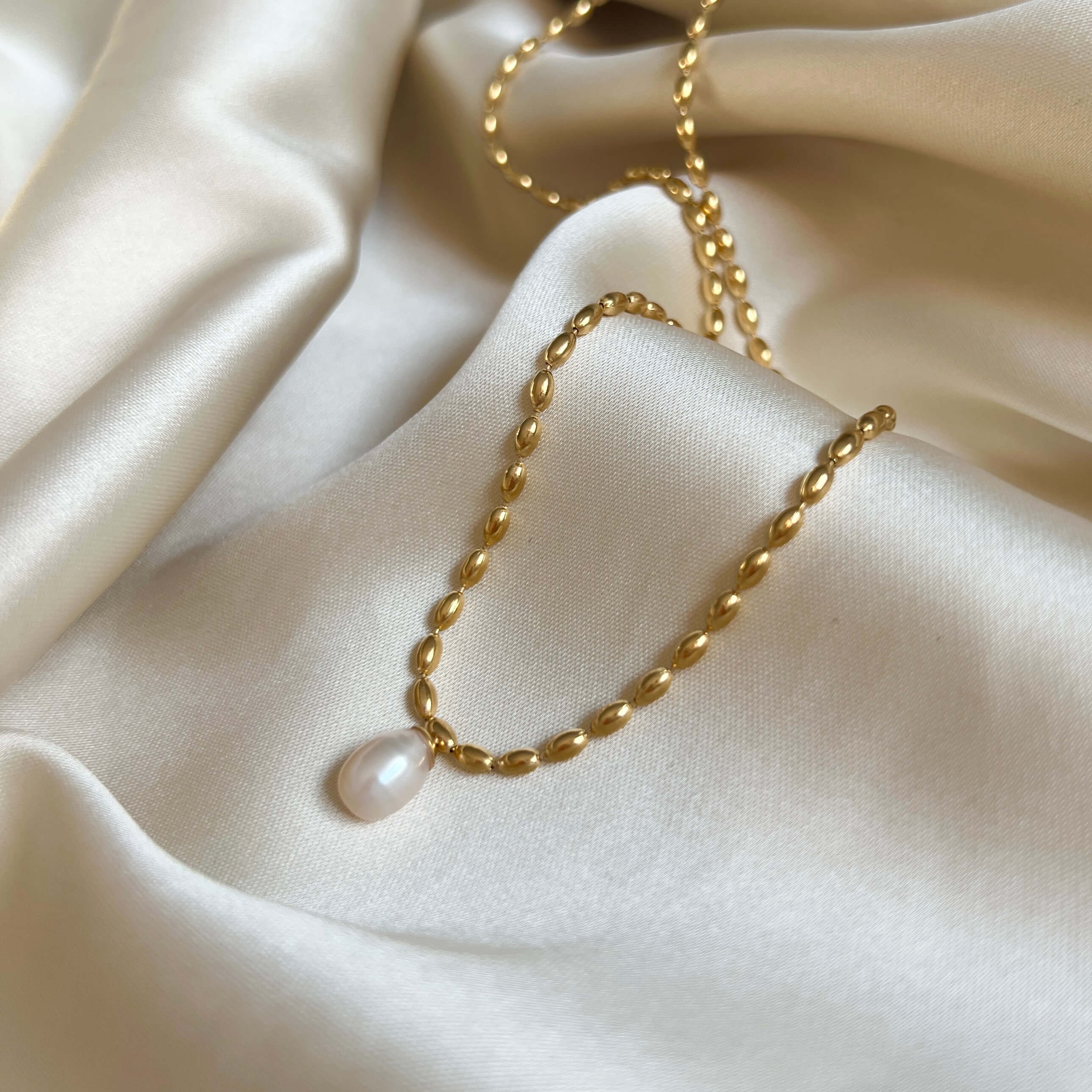 Polly Pearl Necklace från Alora Avenue i närbild – ett vattentåligt halsband i guldfärgat rostfritt stål med droppformad sötvattenspärla, liggande på cremevitt siden. Ett allergivänligt smycke som kombinerar klassisk elegans med modern hållbarhet – perfekt som vardagssmycke eller gåva.