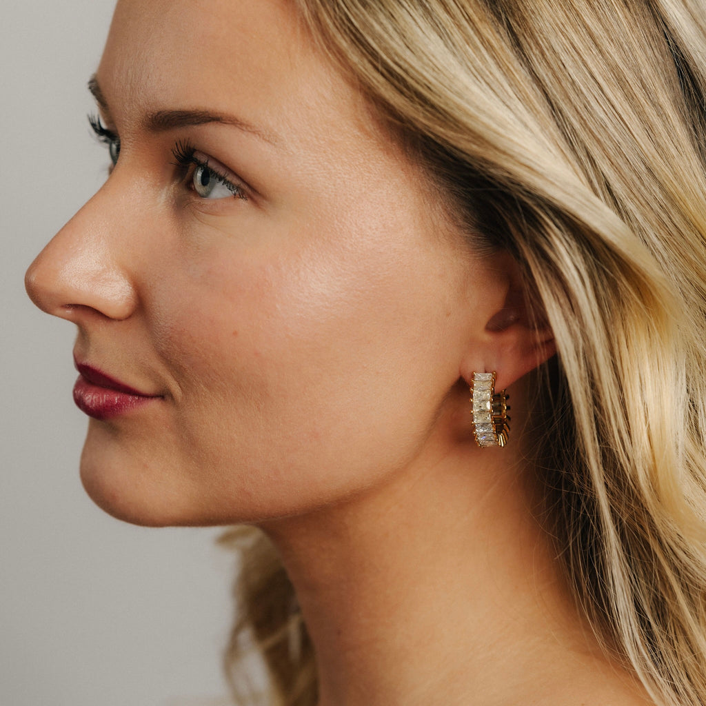 Kvinna bär Sabrina Spark Hoops från Alora Avenue – guldiga örhängen med rektangulära kristaller som glittrar vackert mot huden. Eleganta, vattenresistenta hoops i PVD-pläterat rostfritt stål, perfekta för både vardag och fest.