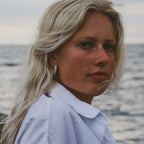 Närbild på modell i vit skjorta vid havet som bär Alora Avenue's glittrande Sabrina Spark Hoops – eleganta guldörhängen perfekta för både vardag och fest. Vattentåliga, allergivänliga smycken med tidlös lyster.