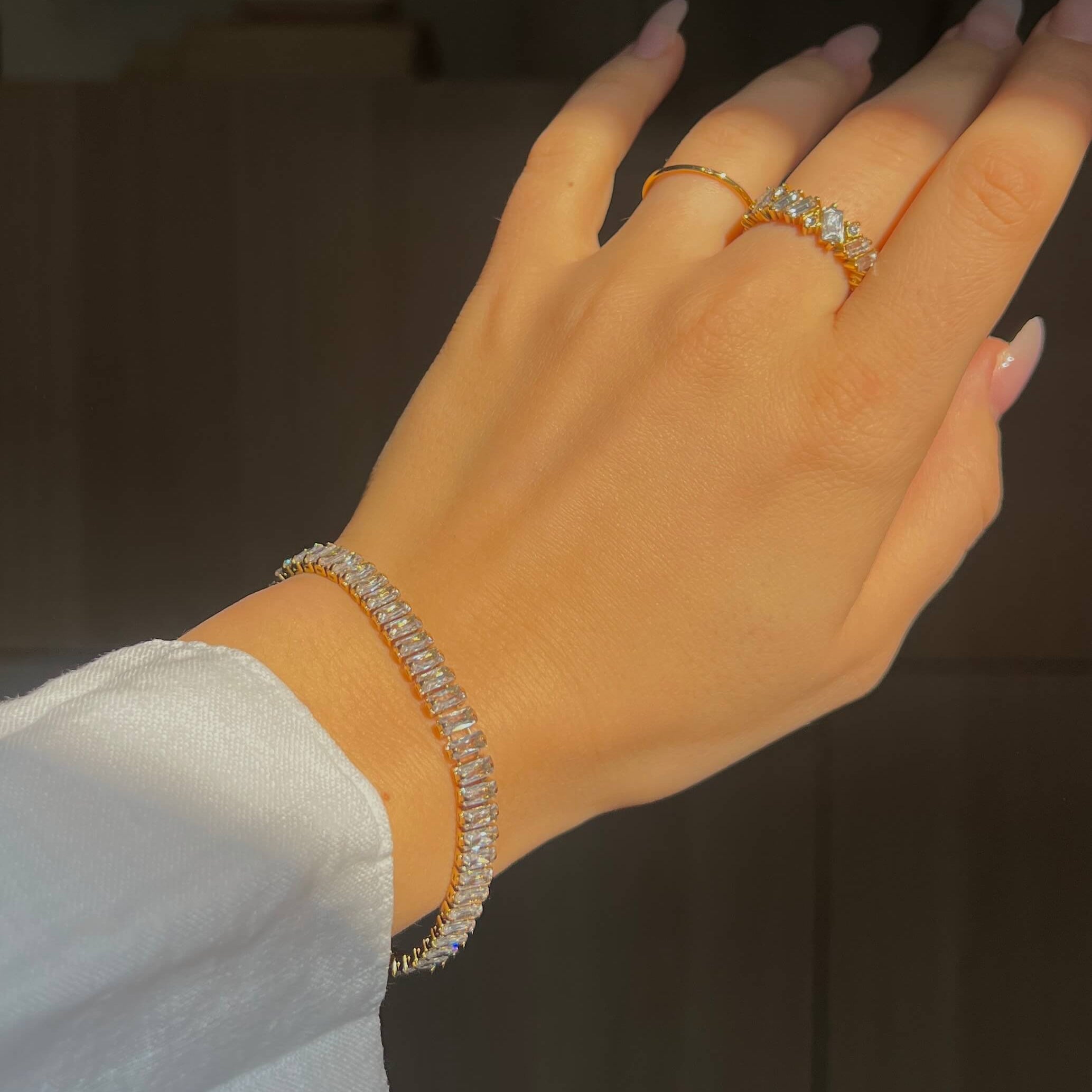 Santiago Sunstone Bracelet bärs på handled i starkt solljus. Armbandet glittrar med baguetteslipade stenar i guldinfattning. Två guldiga ringar syns även på fingrarna.