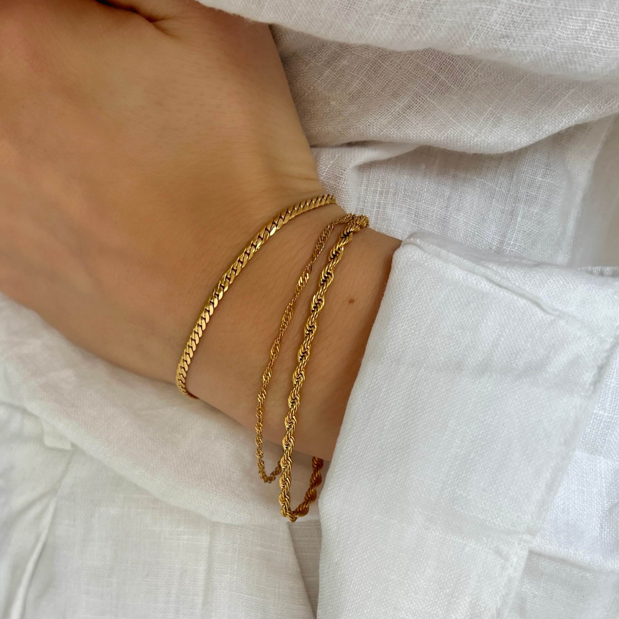 Sia Singapore Bracelet från Alora Avenue – elegant, tvinnat armband i guldfärg. Fotograferat på hand iklädd vit linneskjorta för en stilren och somrig look. Vattentåligt och allergivänligt smycke i rostfritt stål med lyxig finish.