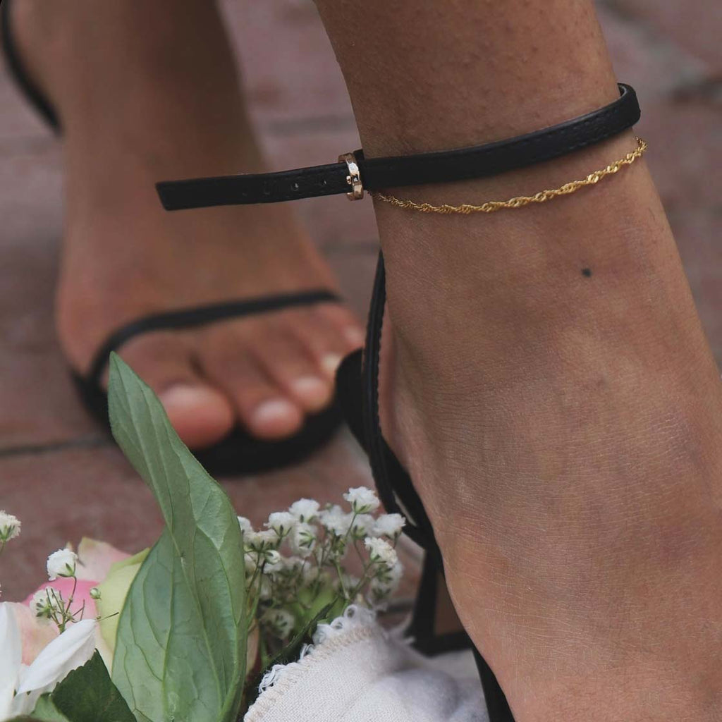 Siri Singapore Anklet från Alora Avenue – tunn, guldfärgad fotlänk med tvinnad design. Bärs tillsammans med svarta klackar och omgiven av blomdetaljer – en elegant och vattentålig accessoar för sommarens alla tillfällen.