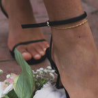 Siri Singapore Anklet från Alora Avenue – tunn, guldfärgad fotlänk med tvinnad design. Bärs tillsammans med svarta klackar och omgiven av blomdetaljer – en elegant och vattentålig accessoar för sommarens alla tillfällen.