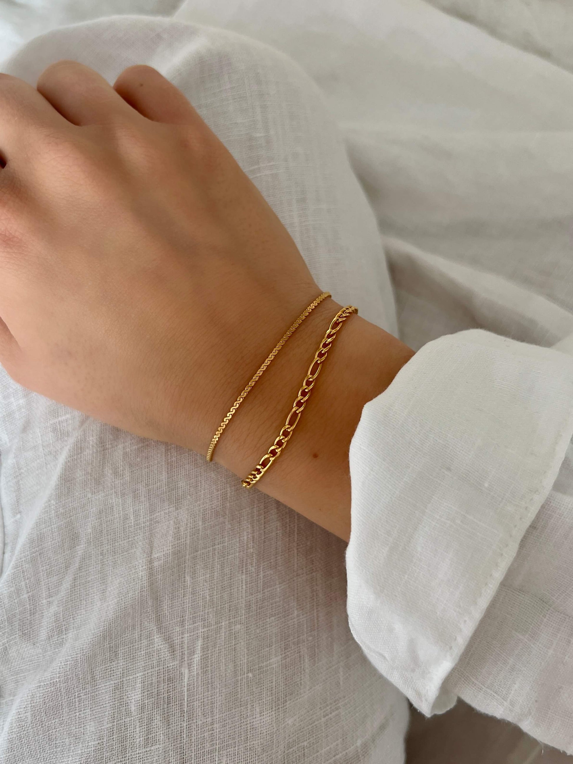 Sonya Saffron Bracelet från Alora Avenue – tunt, minimalistiskt armband i guldfärg. Fotograferat på hand med vit linneskjorta. Vattentåligt, allergivänligt och perfekt för lager på lager-styling.