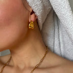 Närbild på ett öra med Stockholm Echo Earrings i glänsande guld – moderna, organiskt formade creoler med en mjuk men kraftfull silhuett. Matchas med en kedjelänkad halskedja och en handduksturban för en avslappnat elegant look.