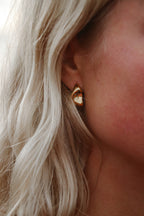 Närbild på ett öra med Stockholm Echo Earrings – glänsande, skulpturala guldcreoler som fångar ljuset. Smycket kontrasterar fint mot det ljusa håret och solkysst hud.