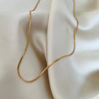 Svea Saffron Necklace från Alora Avenue i närbild – en tunn, elegant guldpläterad kedja i rostfritt stål, fotograferad på cremefärgat tyg. Ett vattentåligt och allergivänligt halsband med diskret vågig struktur – perfekt för lager på lager eller att bära solo varje dag.