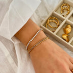 Tilda Tennis Bracelet från Alora Avenue – ett vattentåligt tennisarmband i guldpläterat rostfritt stål med vita gnistrande stenar, här stylat tillsammans med ett chunky guldarmband. Fotograferat mot vit textil med smyckesförvaring i bakgrunden – perfekt för en elegant vardagslook eller som gåva.