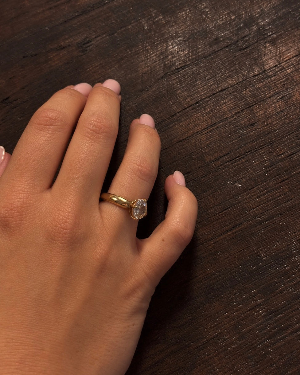 Adele Antique Ring i guld buren på modell mot ett mörkt träbord – antikinspirerad solitärring med en infattad cubic zirconia-sten. Vattentålig, allergivänlig ring i rostfritt stål med 18K guldplätering från Alora Avenue.
