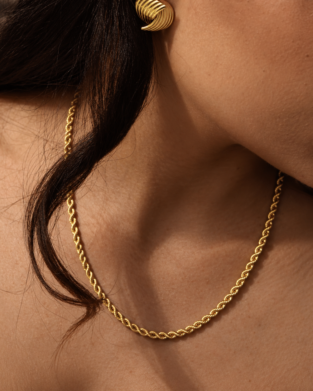 Aloe Annie Necklace i guld på modell – tvinnad repkedja runt halsen, buren till ett räfflat guldörhänge. Vattentåligt och allergivänligt halsband i rostfritt stål med 18K PVD-guldplätering från Alora Avenue.