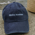 Marinblå keps med vit Alora Avenue-logga broderad framtill – stilren och tidlös merch accessoar, liggandes på sten vid strandväska.
