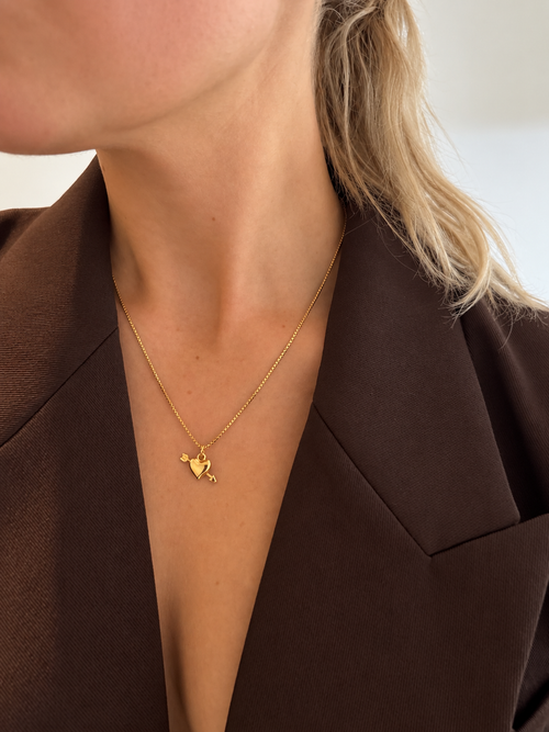 Amora Arrow Necklace – hjärthänge med pil i guld på tunn kedja, buret av modell. Vattentåligt halsband från Alora Avenue.