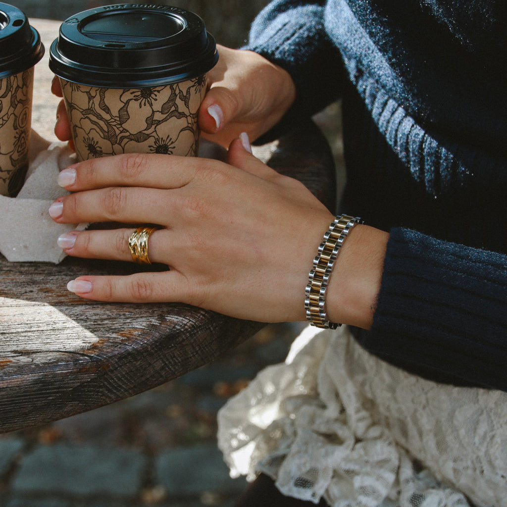 Analog Ava Wristband i guld på handled, stylat med kaffekopp och stickad tröja. Stilrent och vattentåligt armband från Alora Avenue – tidlös design för vardag och fest.