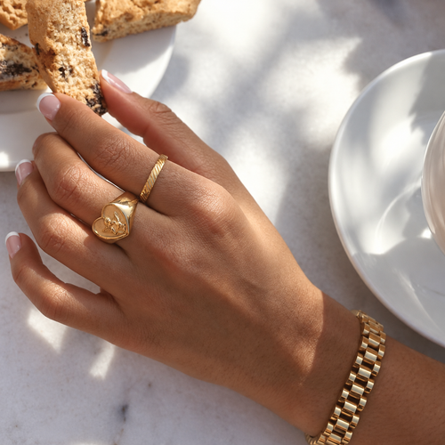 Angelic Love Ring i guld på modell – signetring buren till ett guldarmband i solljus. Vattentålig och allergivänlig ring i rostfritt stål med 18K PVD-guldplätering från Alora Avenue.