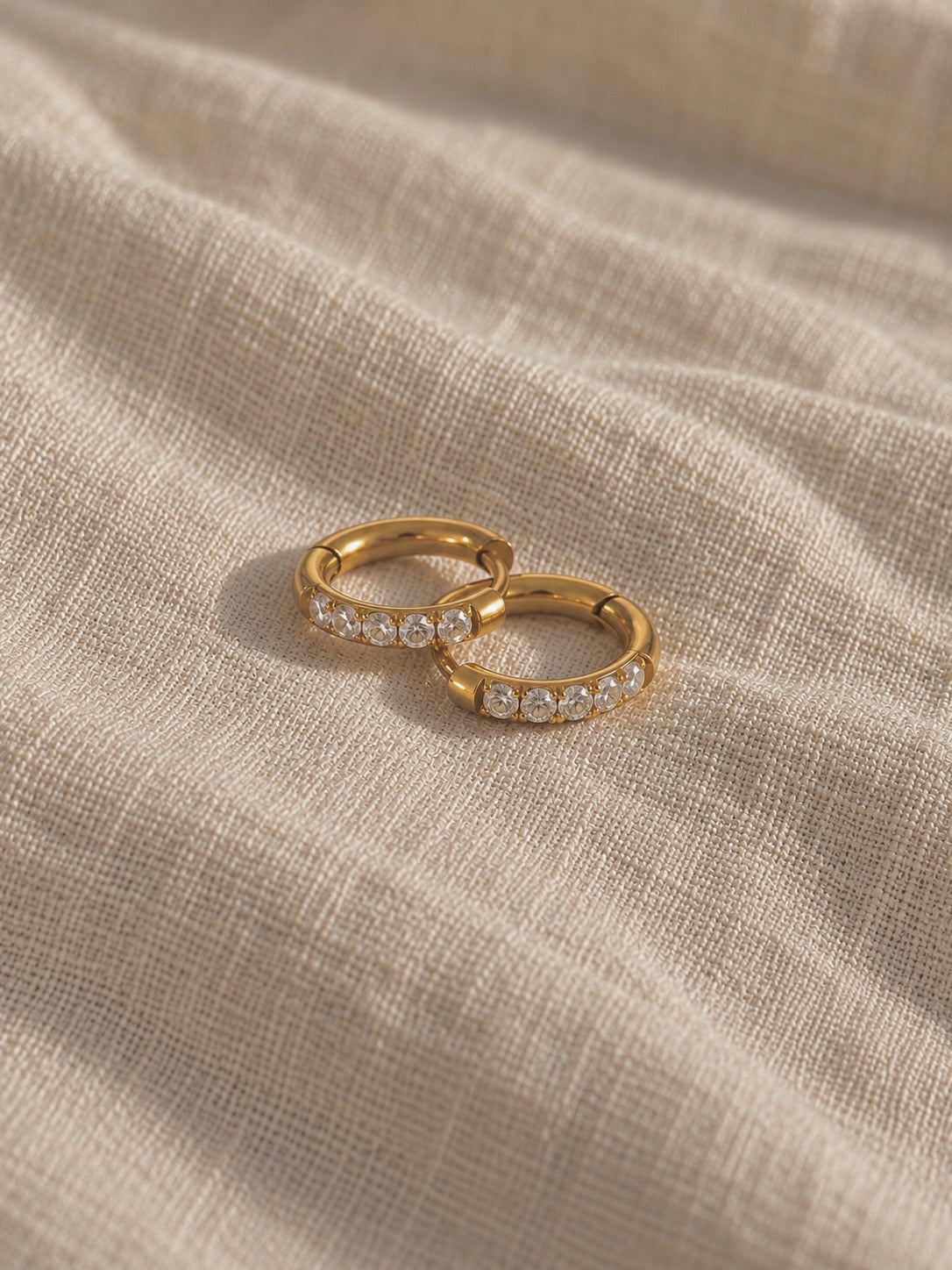 Anni Aura Hoops i guld – ett par pavéhuggies med gnistrande cubic zirconia på linnetyg. Vattentåliga, allergivänliga guldörhängen från Alora Avenue.