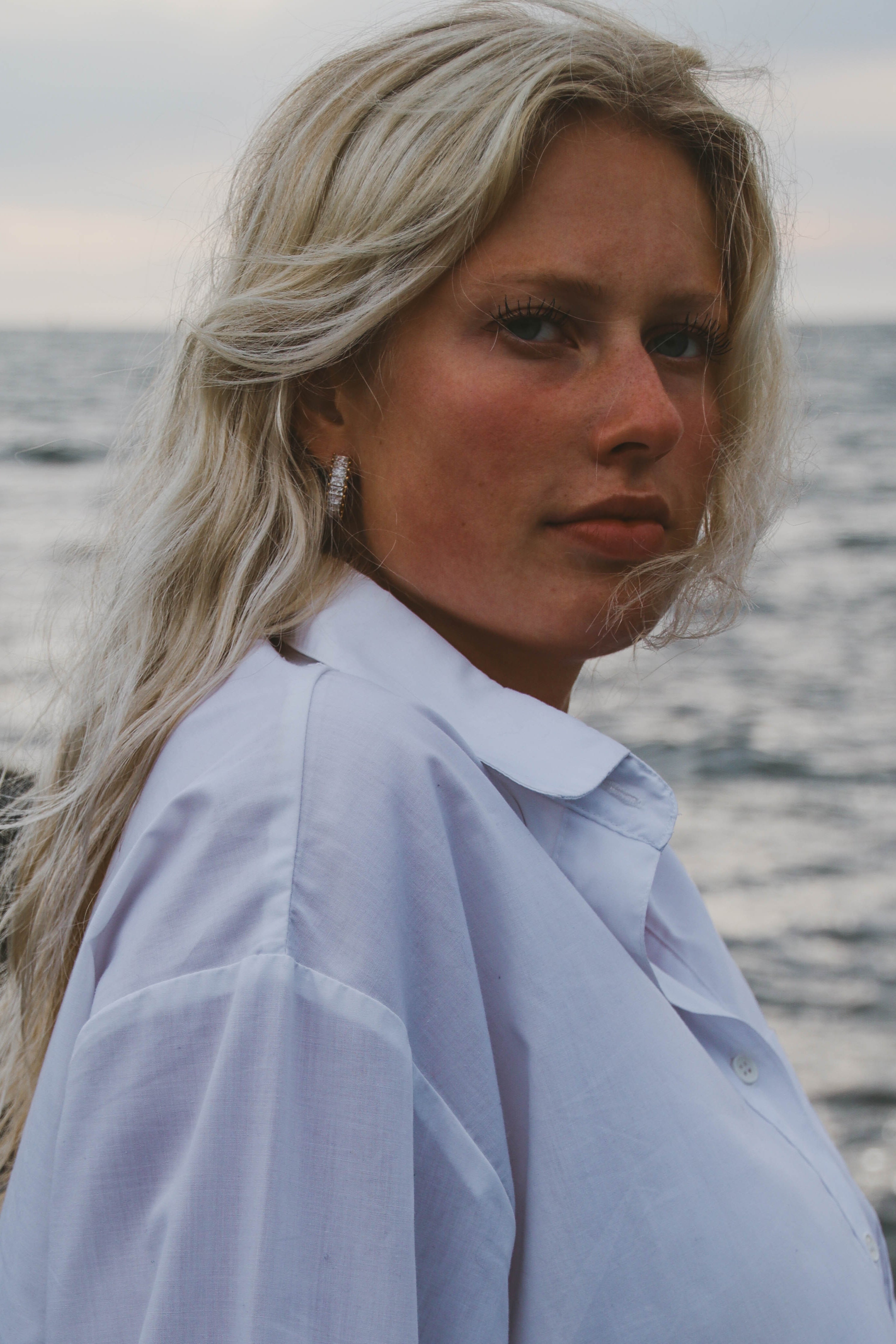Kvinna i vit skjorta vid havet, bär Alora Avenue örhängen – en sensommarlook perfekt för en elegant start på vardagen. Vattentåliga och allergivänliga smycken.