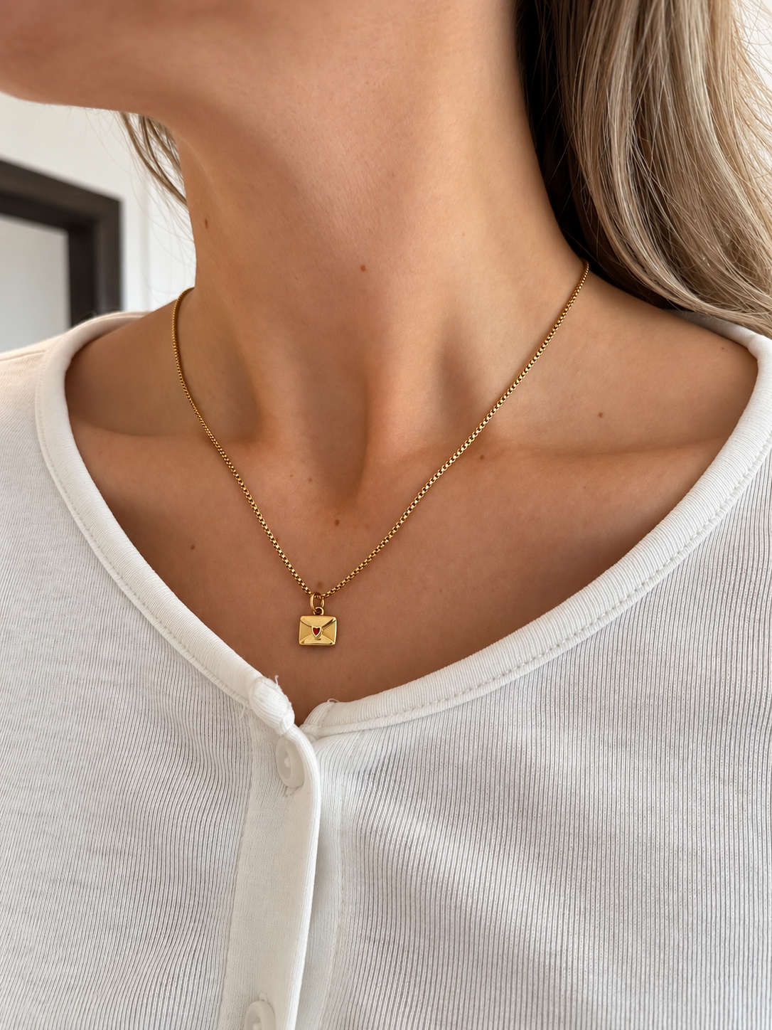 Bisou Letter Necklace – kuvertformat hänge i guld med litet rött hjärta, buret av modell. Vattentåligt halsband från Alora Avenue.