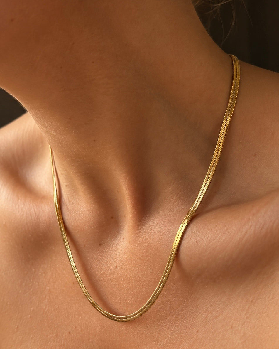Clara Cobra Necklace i guld på modell – platt ormlänkshalsband (snake chain) med blank yta som ligger mjukt mot nyckelbenen. Vattentåligt halsband i rostfritt stål med 18K guldplätering från Alora Avenue.