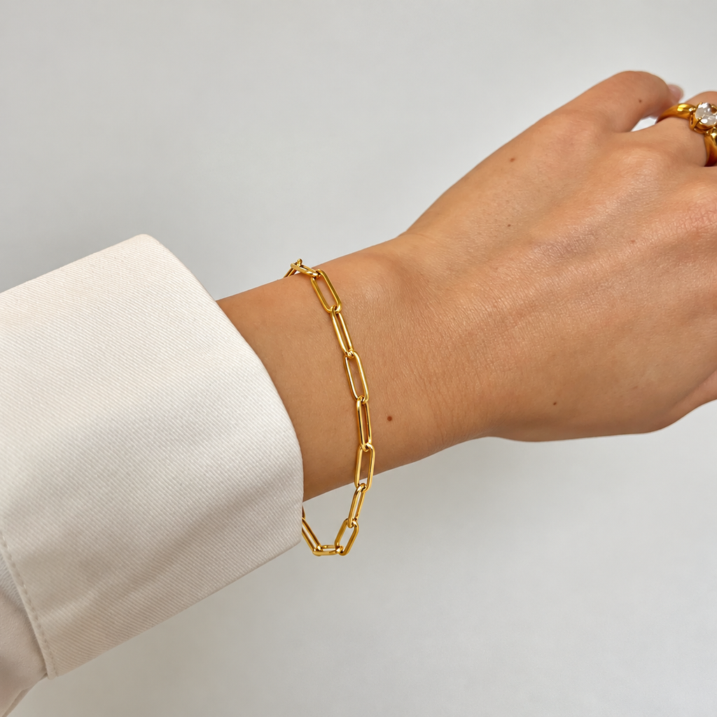 Dawn Clip Bracelet i guld buret på modellens handled, stylat med en guldring. Vattentåligt och allergivänligt armband i rostfritt stål med 18K PVD-guldplätering från Alora Avenue.
