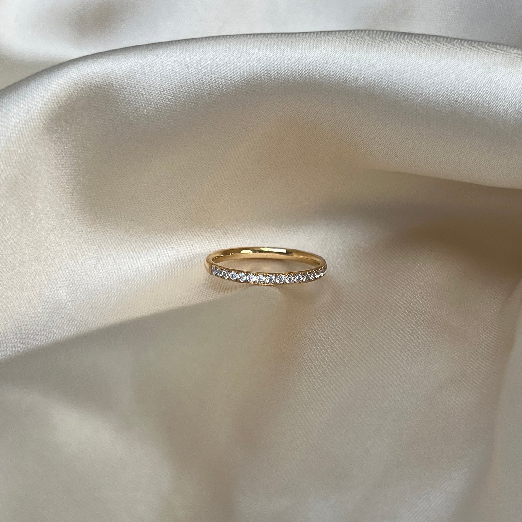 Elara Glow Ring i guld med gnistrande stenar längs halva ringen, fotograferad på ljus sidenbakgrund. Tidlös, vattentålig och allergivänlig ring från Alora Avenue.