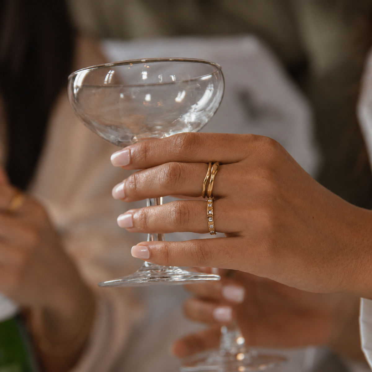 Faye Flow Ring i guld på modell som håller ett coupeglas. Ultrasmal, vattentålig och allergivänlig ring i rostfritt stål med 18K PVD-guldplätering från Alora Avenue.