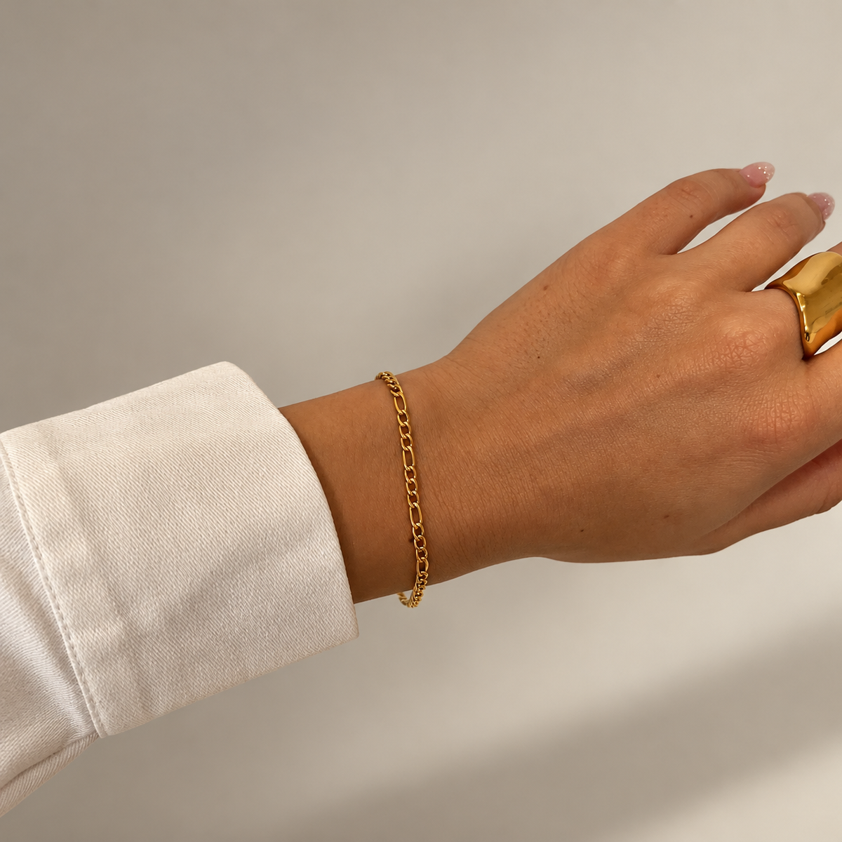 Felix Figaro Bracelet i guld buret på modellens handled till vit skjorta, stylat med en bred guldring. Vattentåligt och allergivänligt armband i rostfritt stål med 18K PVD-guldplätering från Alora Avenue.