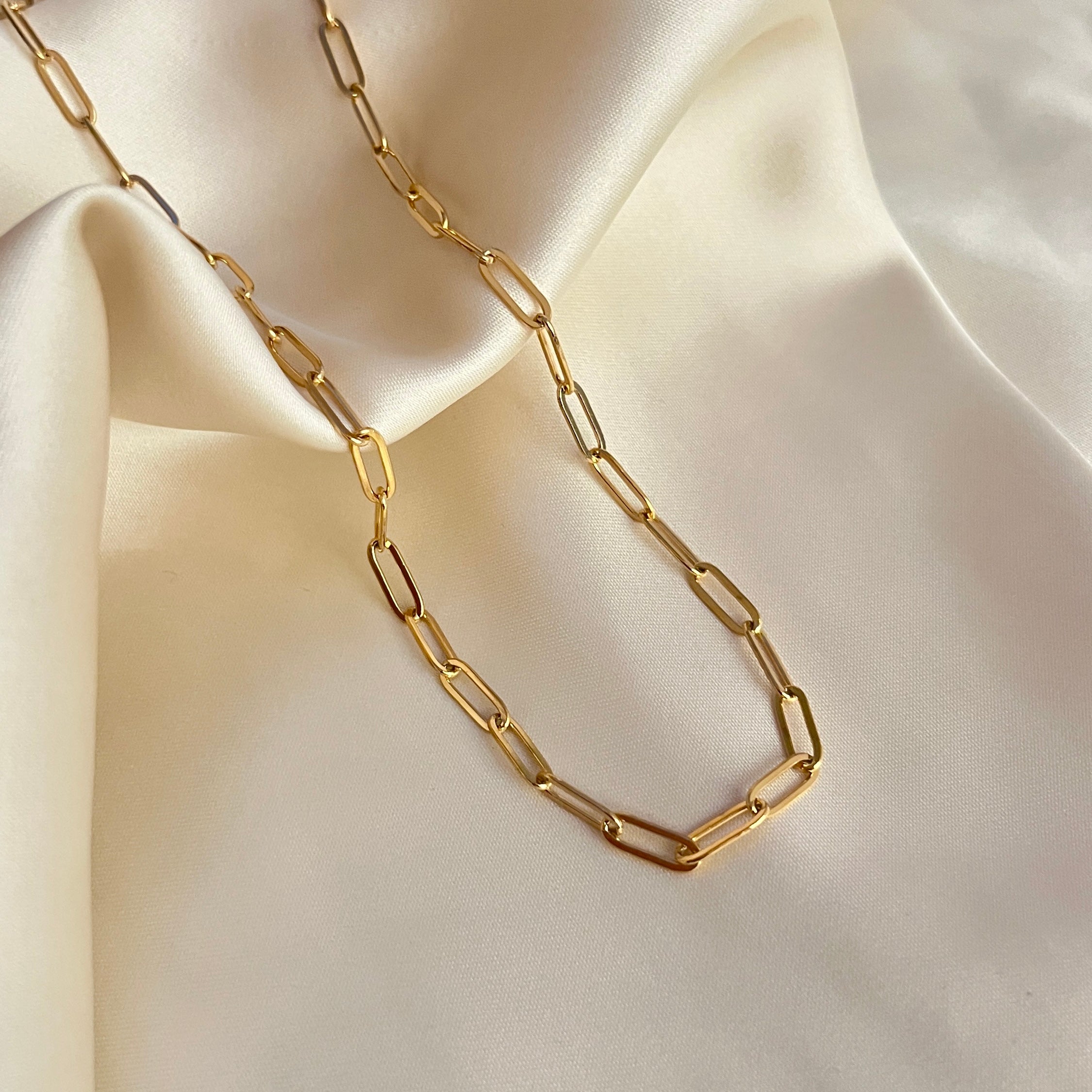 Gala Grove Necklace – stilrent paperclip-halsband i guldpläterat rostfritt stål från Alora Avenue, fotograferat mot champagnefärgat tyg.