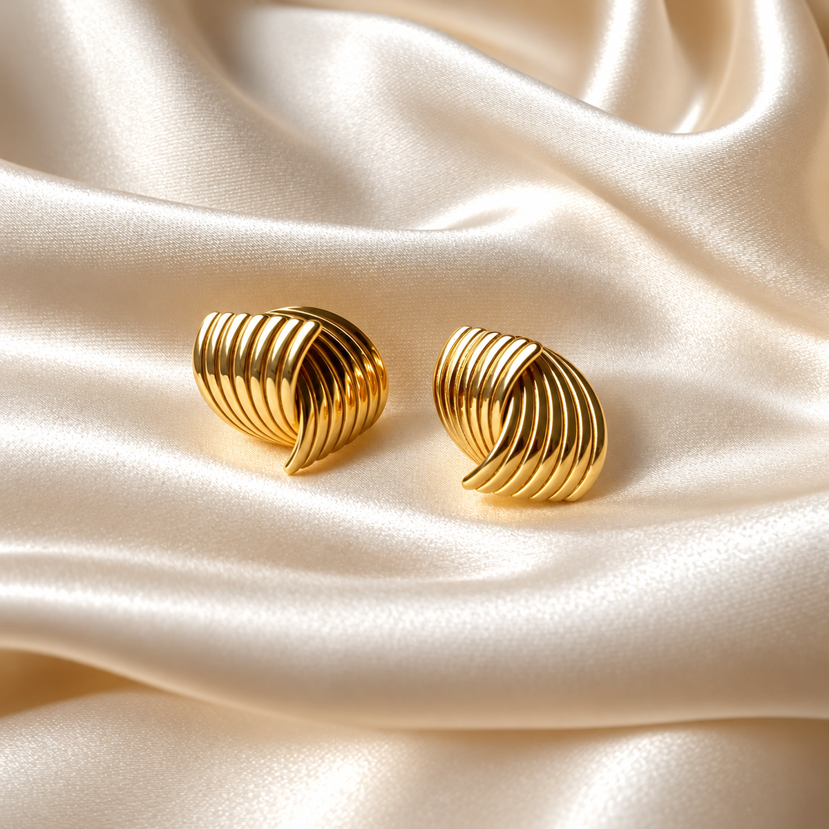 Hanna Huggie Earrings i guld fotograferad på ljust silkestyg. Vattentåligt och allergivänligt smycke i rostfritt stål med 18K PVD-guldplätering från Alora Avenue.