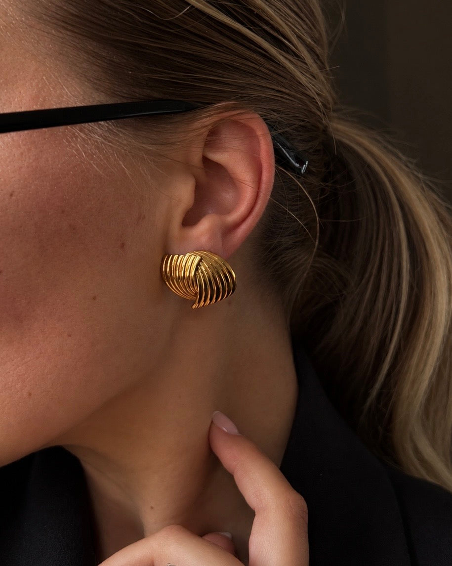 Hanna Huggie Earrings i guld på modell – skulpterade statementörhängen med räfflat, vågigt mönster. Vattentåliga örhängen i rostfritt stål med 18K guldplätering från Alora Avenue.