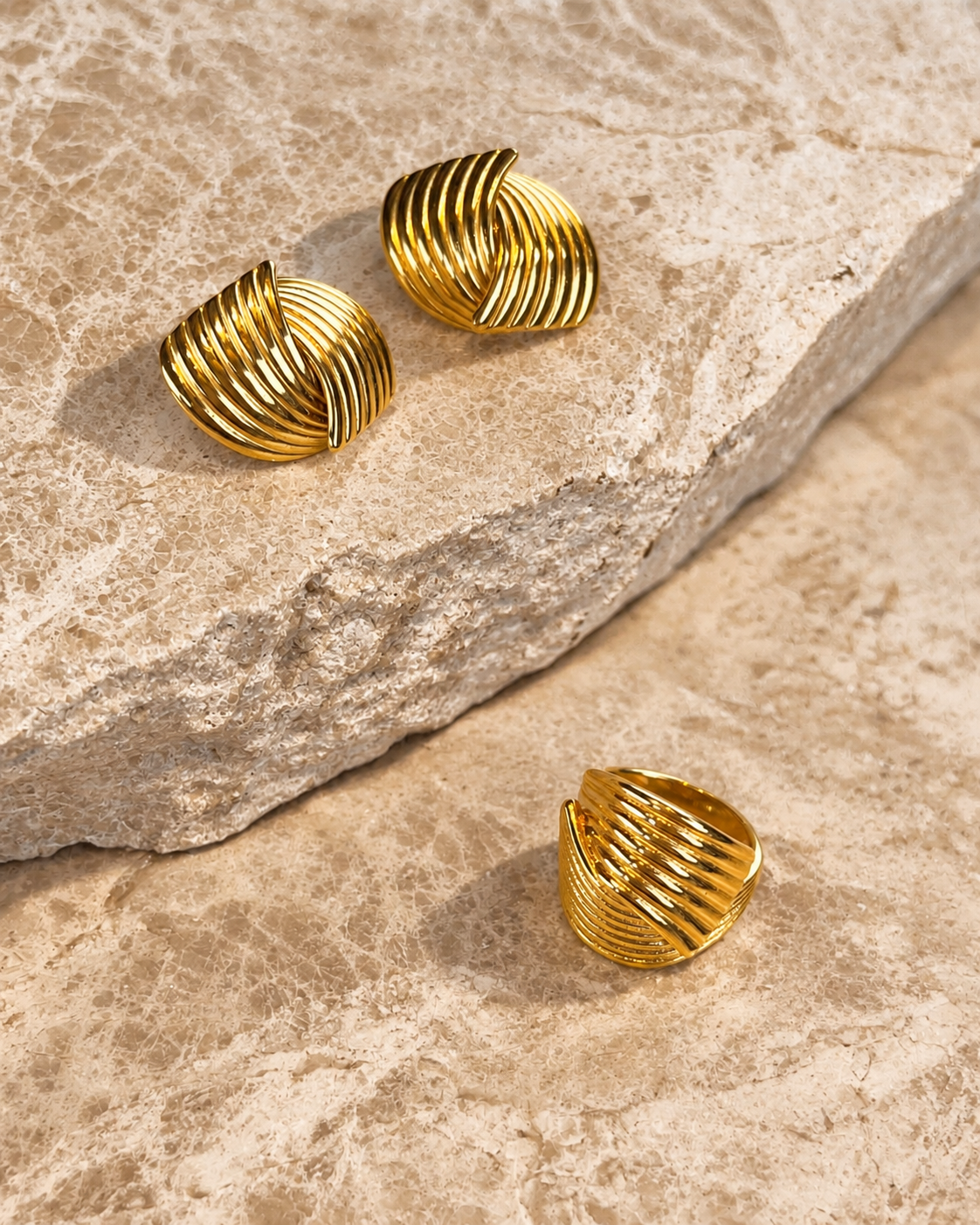 Hanna Huggie Earrings i guld fotograferade på travertin – skulpterade statementörhängen med räfflat, vågigt mönster, visade med matchande Hedda Huggie Ring. Vattentåliga och allergivänliga örhängen i rostfritt stål med 18K PVD-guldplätering från Alora Avenue.