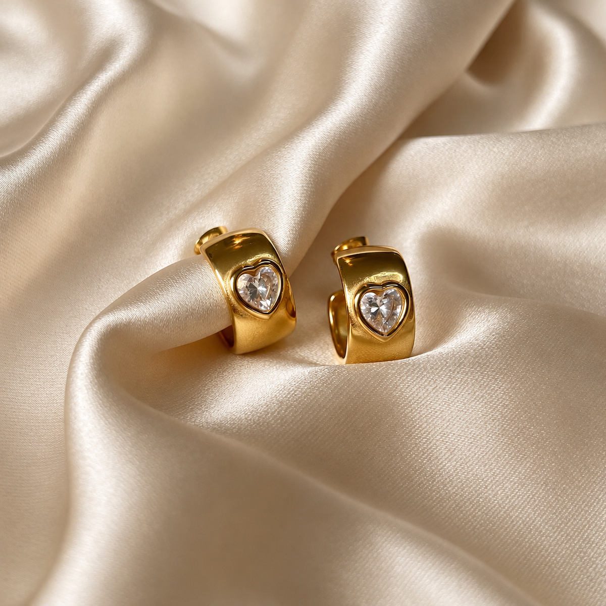 Havanna Heart Earrings i guld fotograferade på ljust silkestyg – breda huggies med en hjärtformad sten. Vattentåliga och allergivänliga örhängen i rostfritt stål med 18K PVD-guldplätering från Alora Avenue.